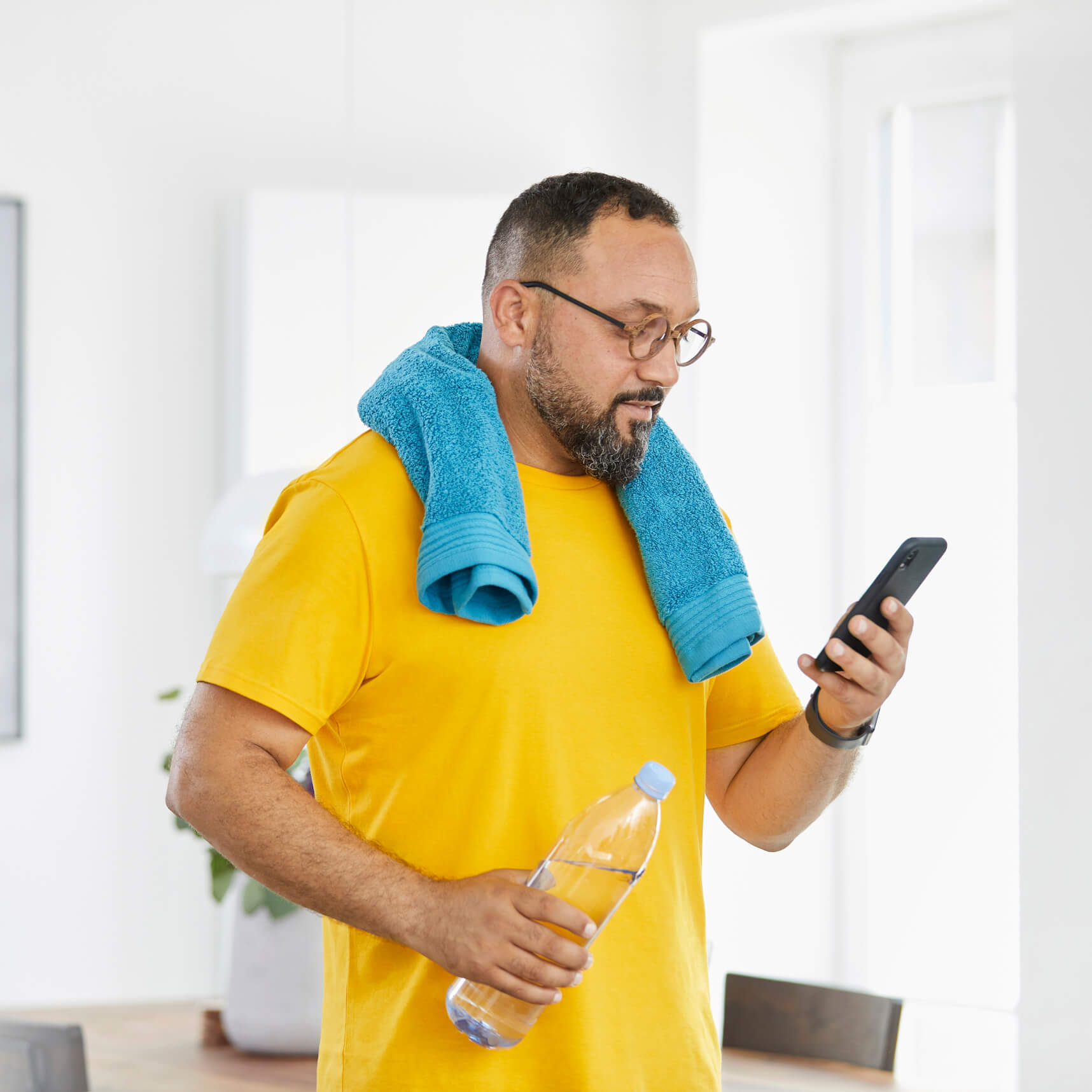 A man wearing a towel around his shoulders and holding a water bottle while looking at a smartphone.